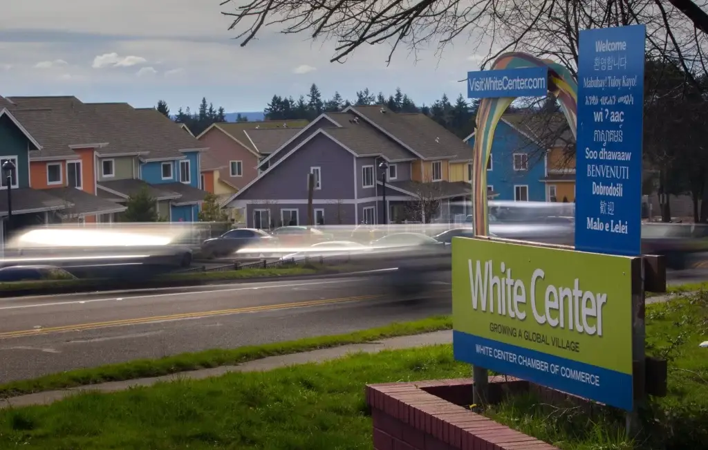 Street view of White Center, WA, with colorful homes, a "Welcome to White Center" sign in multiple languages, and passing cars.