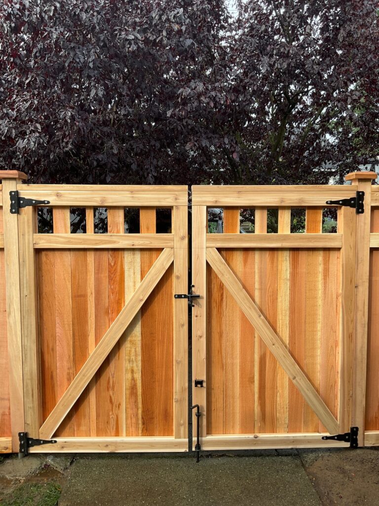 Double wooden gate with a decorative top, reinforced with diagonal braces, secured with metal hinges and latches, against a backdrop of a tree.