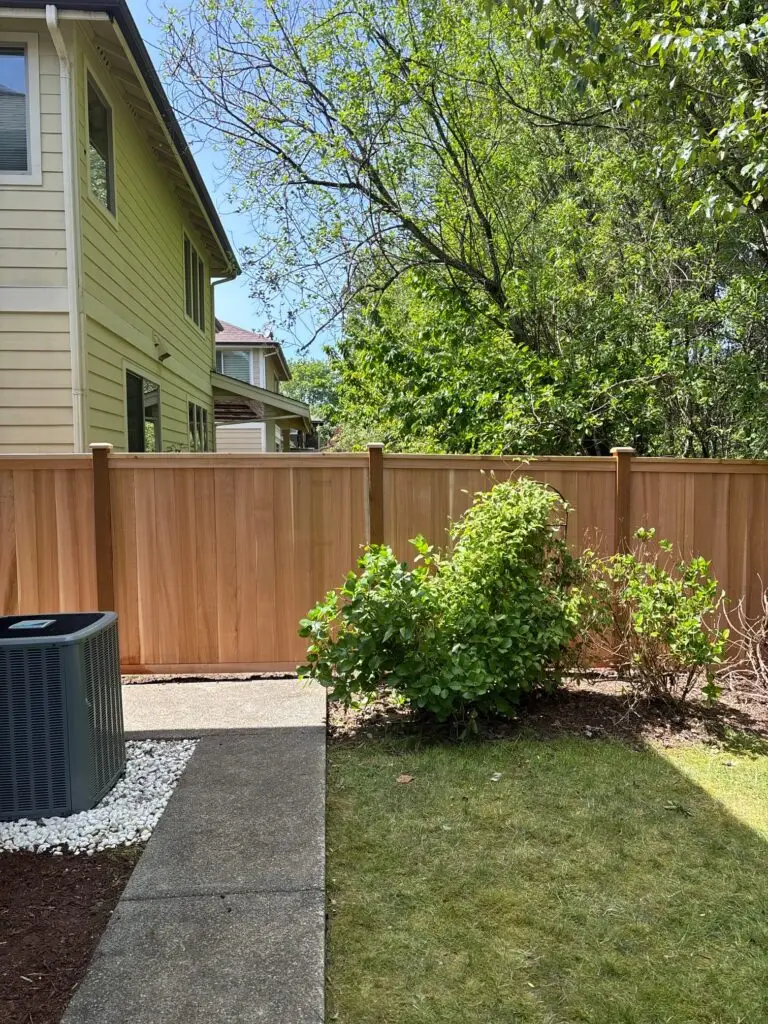 A well-constructed wooden fence in a residential backyard in Seattle with lush greenery and a clean paved path next to the fence. The fence is tall and made from cedar wood, providing privacy.