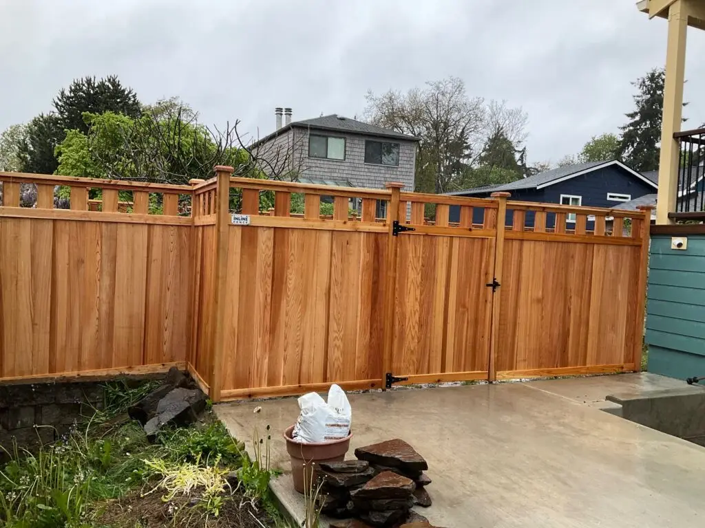 Wooden fence with a decorative top section, showcasing a clean, simple design, accompanied by a matching gate, with natural greenery at the base.