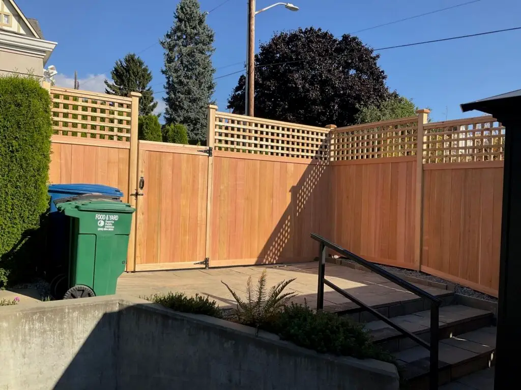 Wooden fence with a decorative lattice top, enclosing a private area with a double gate, against a clear blue sky. The scene also includes a few plants, a staircase, and garbage bins.