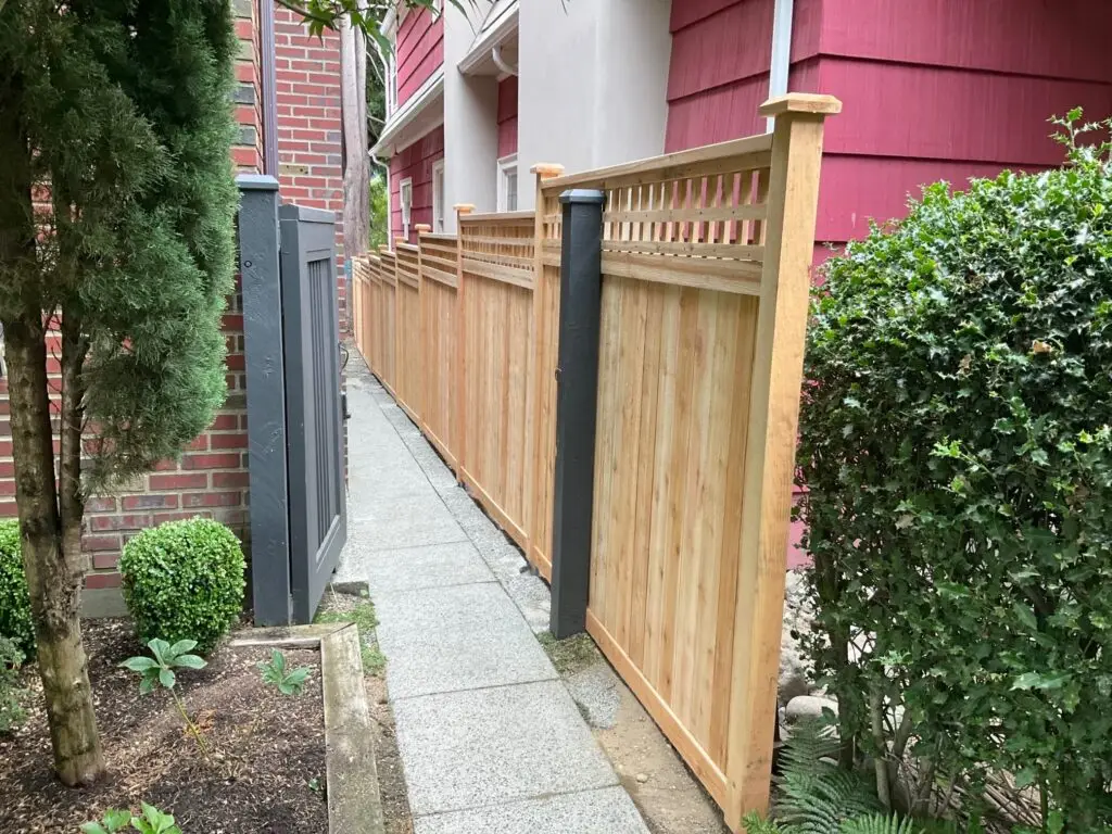 Wooden privacy fence with a modern design, featuring horizontal slats and decorative top, along with a gray gate and surrounding greenery.