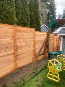 A new horizontal cedar fence separates a backyard play area with swings from a row of tall evergreen trees, designed with careful attention to Horizontal fence height rules.