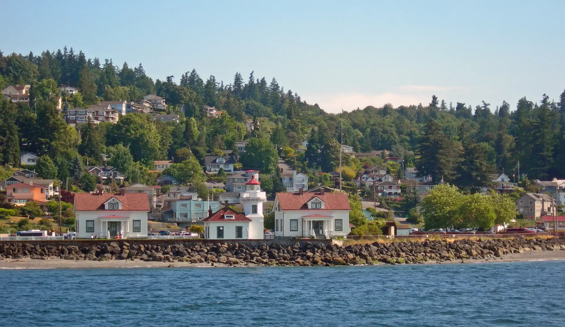 Mukilteo Lighthouse and adjacent white houses with red roofs along the rocky shoreline, with a forested hillside and residential area in the background