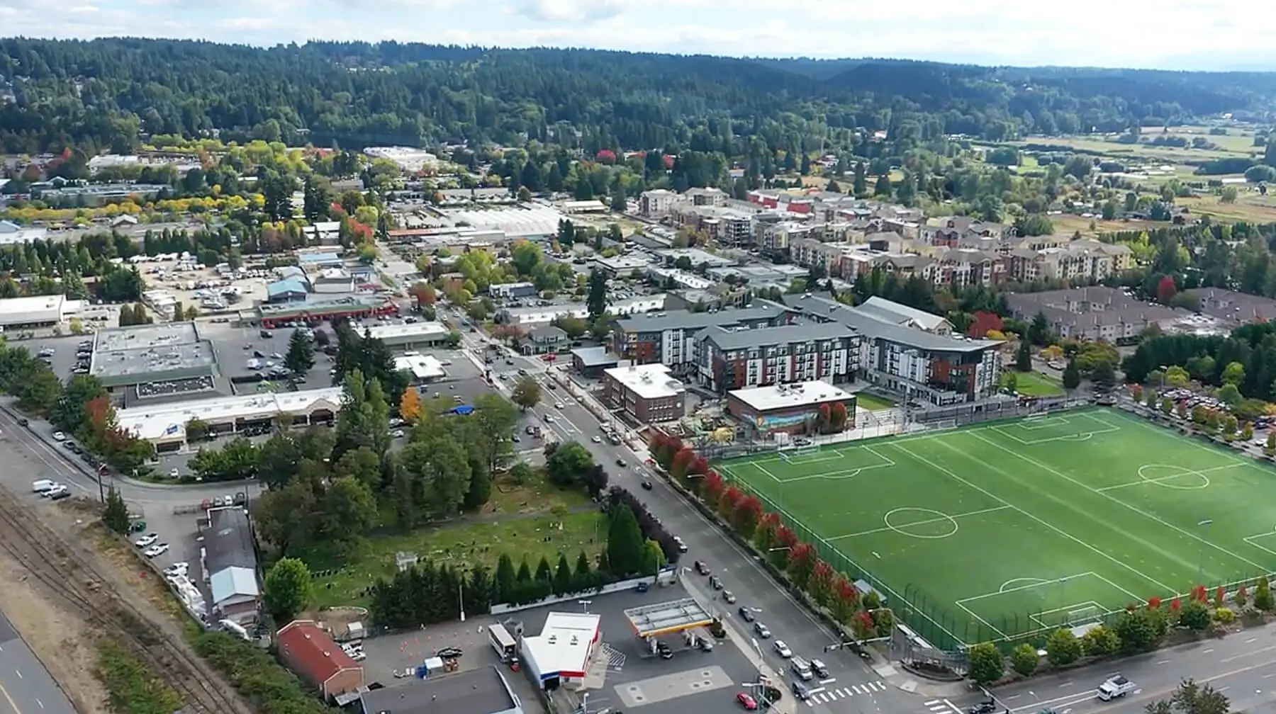 Aerial view of Woodinville, WA, with a soccer field, residential buildings, busy roads, and a forested backdrop under a partly cloudy sky.