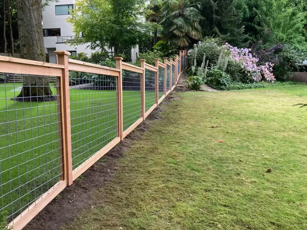 A long, neat row of hog wire fence panels paired with wooden posts, showcasing a well-maintained lawn and lush garden.