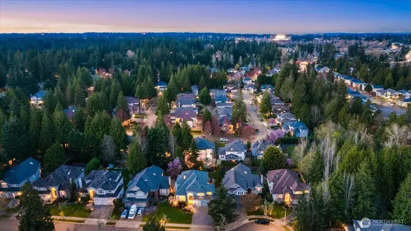 Aerial view of a suburban neighborhood in Mill Creek, WA, with houses nestled among dense trees, illuminated at dusk with a city skyline in the distance.