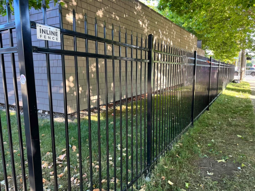 A black ornamental iron fence with an Inline Fence sign, bordered by green grass and a building wall.
