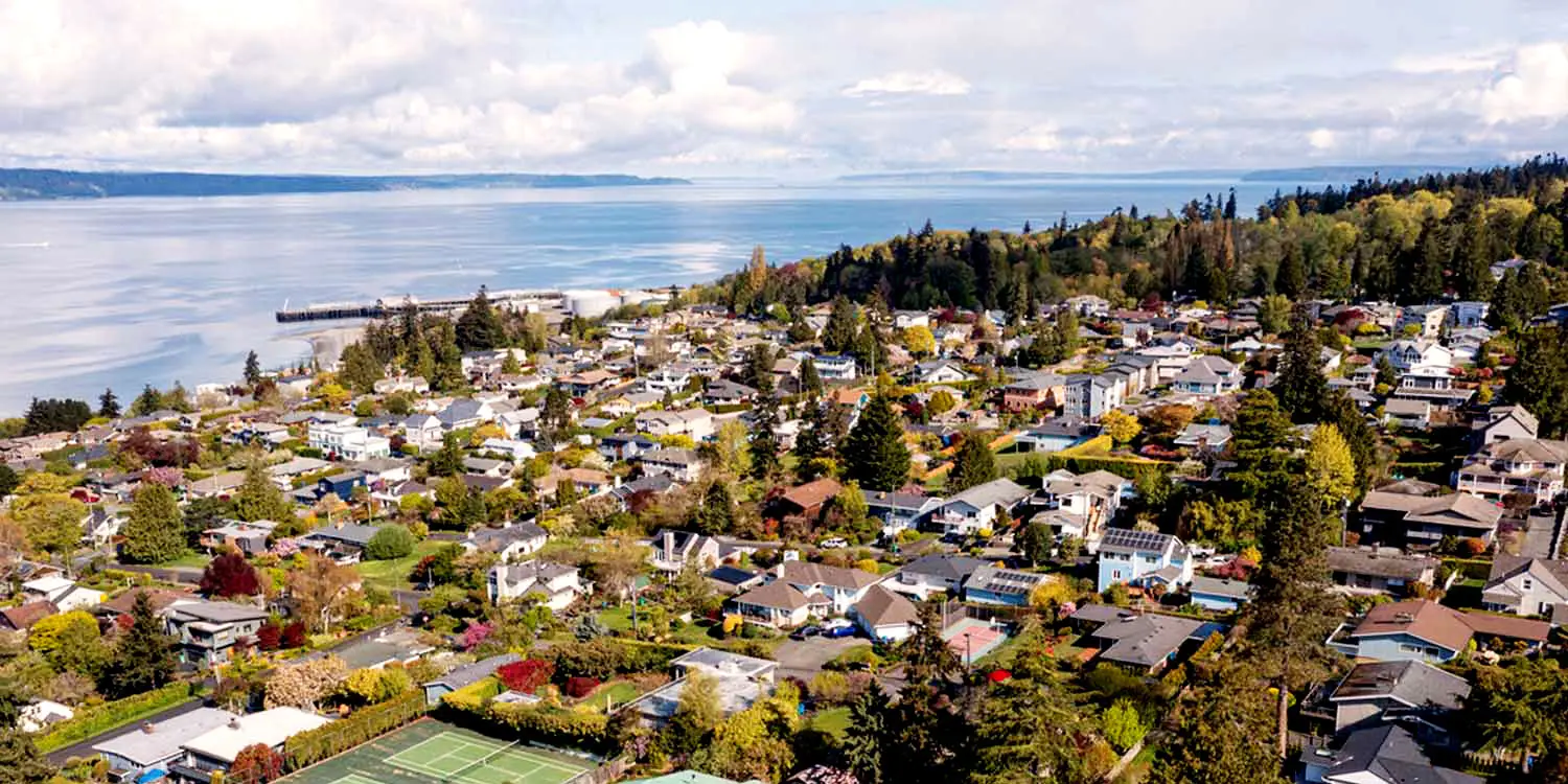 Aerial view of a Shoreline, WA neighborhood with houses, green trees, and a tennis court, near a waterfront with a distant pier and forested hills.