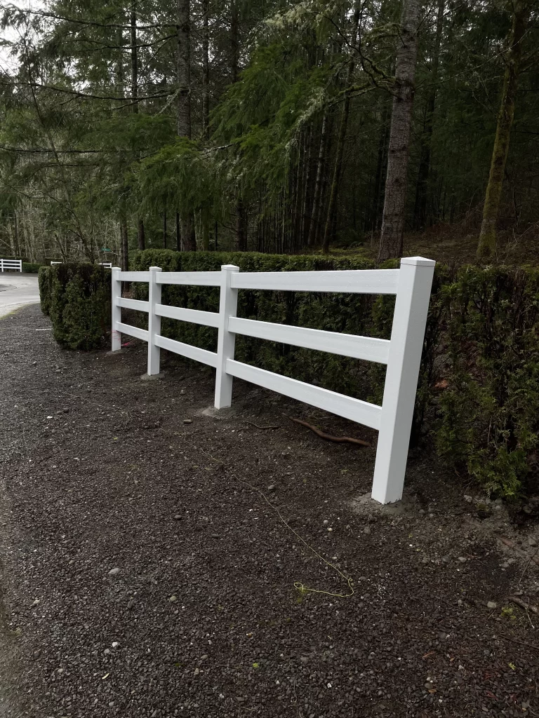 A white vinyl fence with horizontal rails, running along a gravel road with evergreen hedges, reflecting the vinyl fence cost in Seattle.