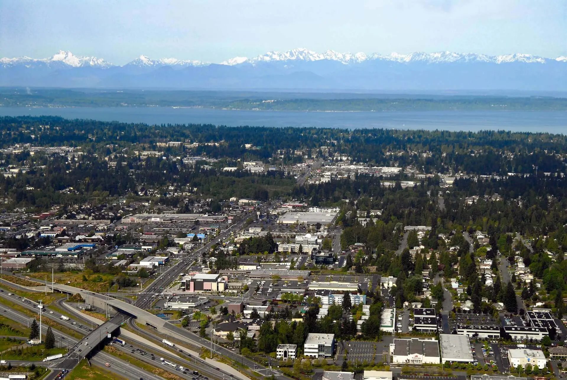Aerial view of a city with dense buildings, trees, highways, and a large body of water with snow-capped mountains in the background under a clear sky.